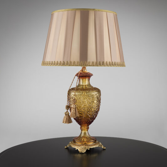 An ornately designed golden table lamp with an intricate base and urn-like shape, reminiscent of the Classic Style Faceted Amber Crystal Table Lamp. It features a beige pleated lampshade with golden trim, adorned with hanging tassels. The lamp is placed on a black circular table against a plain gray background.