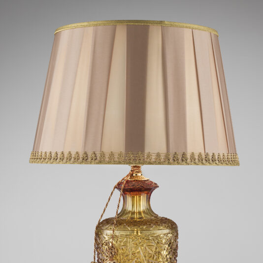 Classic Style Faceted Amber Crystal Table Lamp The Classic Style Faceted Amber Crystal Table Lamp boasts an ornate, clear and gold-toned base with intricate patterns. It features a light brown, shiny fabric lampshade trimmed in gold and an elegant braided cord. The plain, gray background accentuates its timeless design.