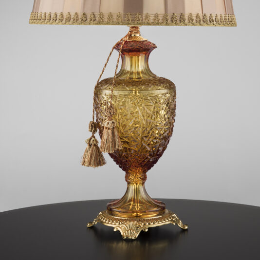 Classic Style Faceted Amber Crystal Table Lamp The Classic Style Faceted Amber Crystal Table Lamp is an ornate, gold-toned table lamp featuring an intricately patterned glass base with red accents at the top and bottom. At its center, there's a faceted amber crystal, complemented by a beige pleated lampshade adorned with decorative tassels. This elegant lamp rests on a round, black surface.
