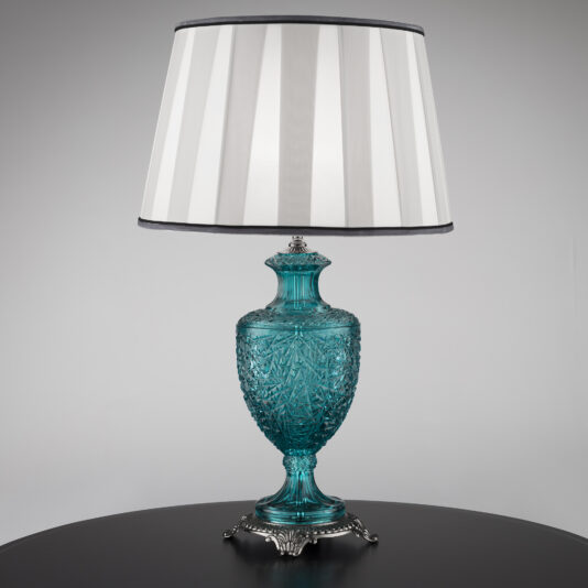A Classic Style Faceted Turquoise Crystal Table Lamp boasting a faceted turquoise crystal base and an elaborate silver stand, crowned with a large white lampshade decorated with vertical silver stripes, set against a plain background.