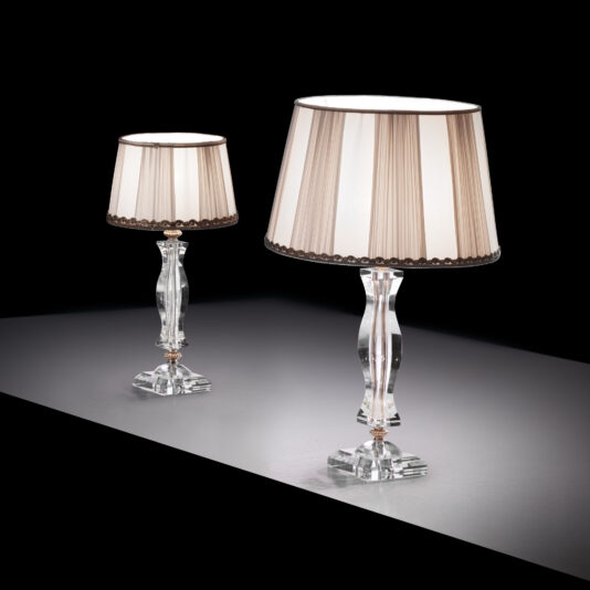 Two Clear Glass Table Lamps With Pleated Shades, featuring white fabric accented with dark trim, are placed on a glossy, dark surface. One lamp is slightly taller than the other, creating a staggered, sophisticated look against the dark background.