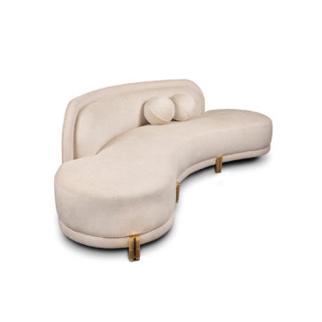 A modern, beige, curved sectional sofa with two cylindrical pillows placed on the left side. The sofa features gold metallic legs and a unique, sculptural design with a high backrest on one end and a low, sweeping contour on the other.