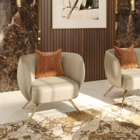 A modern living room features a Contemporary Leather Tub Armchair paired with beige armchairs with gold legs, each adorned with an orange patterned cushion. The backdrop showcases dark marble walls, wooden paneling, and a large abstract artwork. Sunlight brightens the space, highlighting a plush, earth-toned carpet.