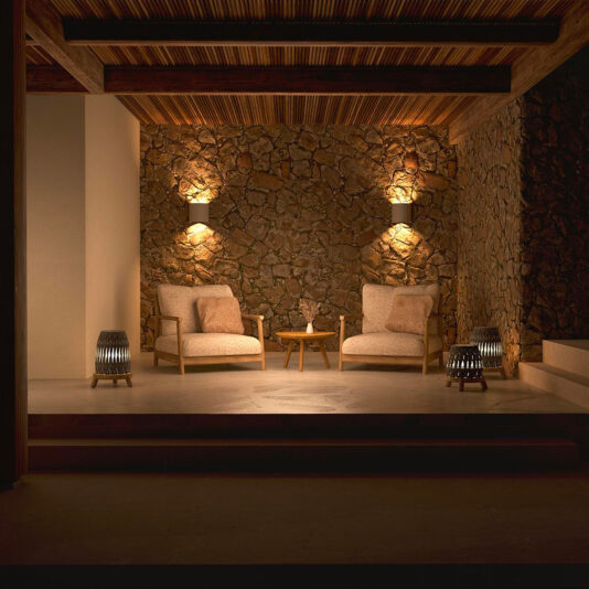 A cozy, dimly-lit seating area featuring two Contemporary Outdoor Deep Teak Armchairs and a small round wooden table in between. The space is surrounded by rustic stone walls and wooden ceiling beams, with wall-mounted and lantern lights creating a warm, inviting ambiance.