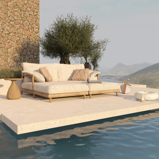 A cozy outdoor lounge area is set on a stone patio by a pool, featuring the Contemporary Outdoor Double Teak Daybed adorned with patterned pillows, complemented by a small round side table and an olive tree in the background. Scenic hills and a clear sky form the serene backdrop.