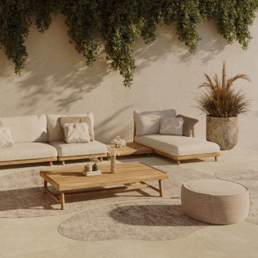 Contemporary Outdoor Teak Garden Set An outdoor patio area showcasing the Contemporary Outdoor Teak Garden Set with beige sofas and cushions, accompanied by a wooden coffee table decorated with various items. A round beige ottoman complements the seating arrangement, while a large potted plant featuring tall, wispy grass is positioned nearby. Leafy green vines dangle gracefully from the top of the beige wall.