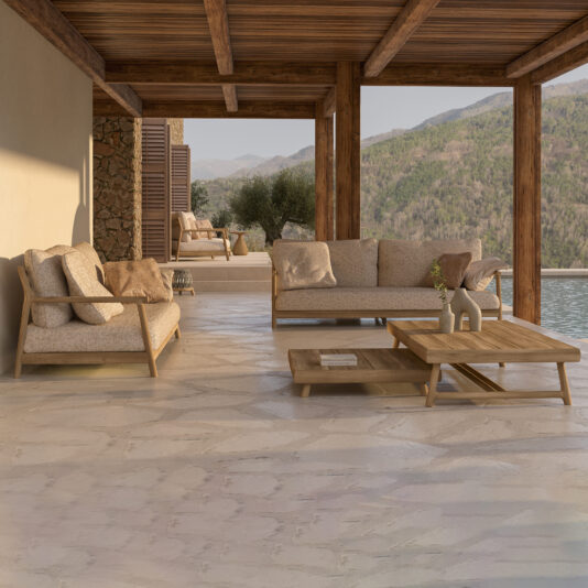 A scenic outdoor lounge area with wooden beams and plush beige furniture, featuring a Contemporary Outdoor Teak Sofa, an additional sofa, and a low coffee table. The space overlooks a mountainous landscape under a clear sky, with sunlight casting a warm glow over the stone floor.