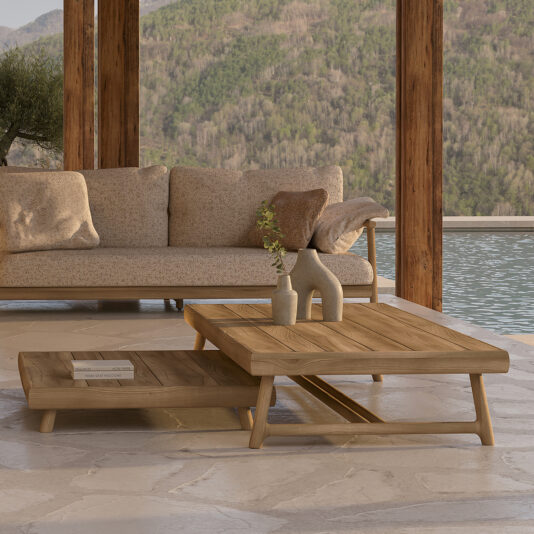 An outdoor seating area featuring a beige sofa adorned with brown and beige cushions. In front, there is one of our Contemporary Slatted Teak Outdoor Coffee Tables, showcasing a sophisticated two-tiered design. A ceramic vase filled with greenery and a book rest gracefully on the table. Behind it lies a pool set against a picturesque backdrop of lush forest scenery.