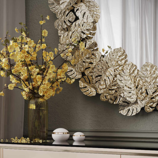 A decorative interior featuring the **Contemporary Tropical Leaf Round Mirror** framed with metallic, leaf-shaped accents and a vase filled with vibrant yellow flowers. The arrangement sits atop a sleek, black surface, lending a luxurious and artistic touch to the setting.