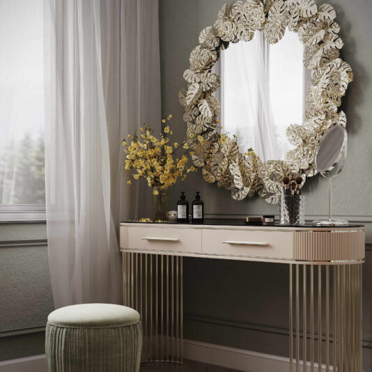 A contemporary round mirror with a decorative tropical leaf frame, known as the Contemporary Tropical Leaf Round Mirror, elegantly hangs above a stylish dressing table. On the table are bottles, a small mirror, and makeup brushes organized in a holder. A vase filled with yellow flowers is placed beside these items. Under the table sits a green cushioned stool placed neatly in its spot.
