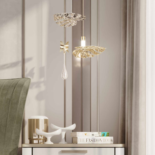 A stylish interior scene featuring the "Glass Pendant Light Inspired By Nature" with artistic designs. A green chair sits next to a light-colored nightstand, which holds a decorative sculpture, a candle holder, and a few books, including "The Fashion Book." Elegant curtains frame the composition.
