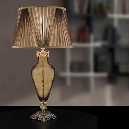 The Hand Blown Brown Glass Table Lamp, adorned with an elegant pleated lampshade in shades of brown, sits on a black surface. The background reveals a blurred interior space with shelves and indistinct objects.