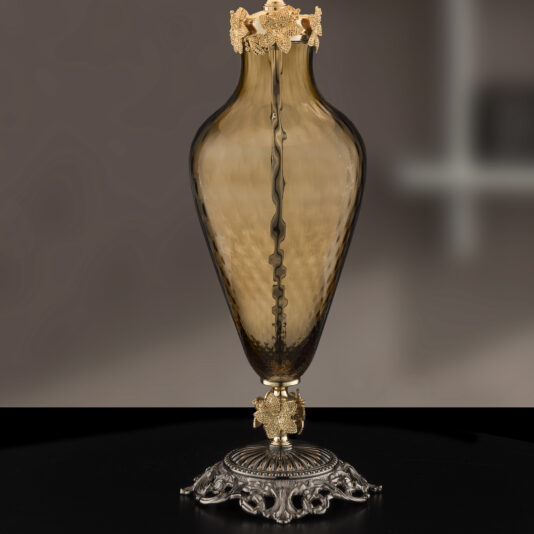 A decorative Hand Blown Brown Glass Table Lamp with a slender, elongated shape. The lamp features intricate gold floral embellishments around the neck and base. It stands on an ornate, silver-colored metal base with detailed, flowing designs. The background is blurred.