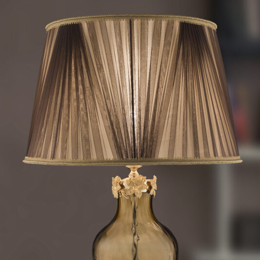 A close-up image of the Hand Blown Brown Glass Table Lamp features a pleated brown lampshade with gold trim. The lamp has an ornate gold base with intricate detailing and sits against a blurred, neutral background.