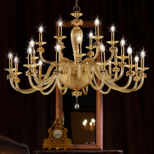 An ornate, gold, multi-tiered Hand Blown Glass Chandelier With Flowers and numerous lit candles hangs in an elegant room. Behind it, there's a framed mirror and an antique clock on a wooden mantle. The background has a dark, rich tone that accentuates the chandelier's bright glow.