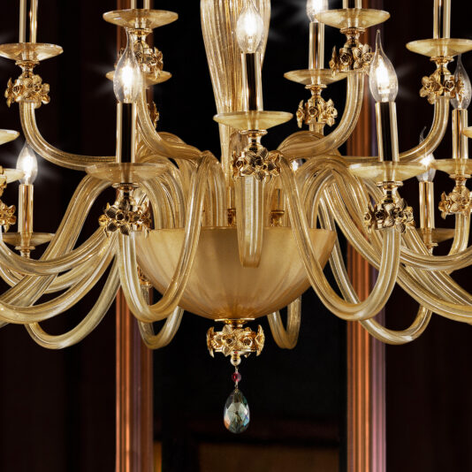 The Hand Blown Glass Chandelier With Flowers is a stunning ornate fixture featuring multiple arms adorned with lit candle-like bulbs. This golden chandelier showcases intricate details and decorative elements, including a dangling drop-shaped crystal. Set against a dark background, it highlights its elegance and intricacy beautifully.