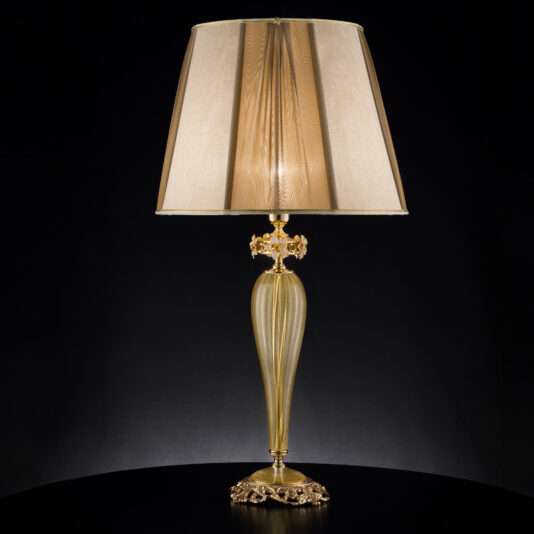 The Hand Blown Glass Table Lamp With Flowers stands tall and elegant on a black surface against a dark background. It features an intricately designed gold base that resembles hand blown glass with delicate flowers, paired with a beige lampshade boasting a subtle striped texture. This exquisite lamp radiates a soft, warm light.