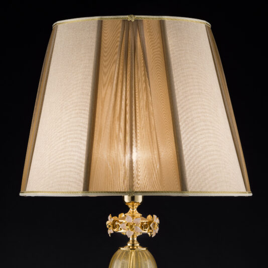 A classic table lamp, featuring a beige lampshade and an ornate golden base adorned with decorative floral elements, is illuminated against a black background, capturing the intricate beauty of the Hand Blown Glass Table Lamp With Flowers.