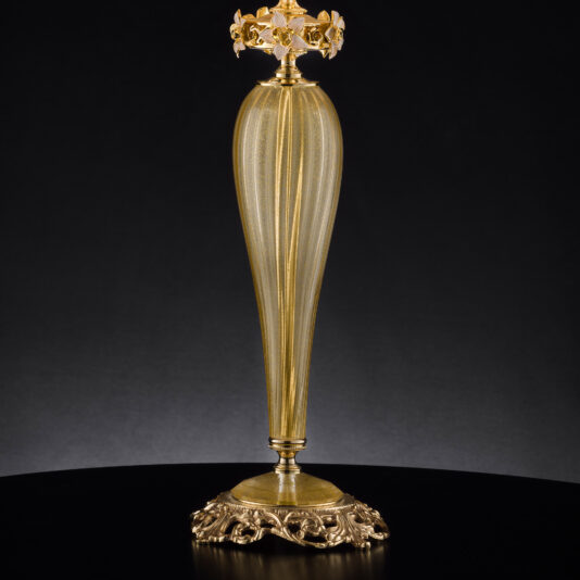 A tall, ornate candlestick with a ribbed, gold-toned body and an intricate base and top. The base features elaborate decorative patterns, while the top is designed to hold a candle with a decorative holder resembling floral or leaf motifs, reminiscent of the Hand Blown Glass Table Lamp With Flowers. The background is dark.