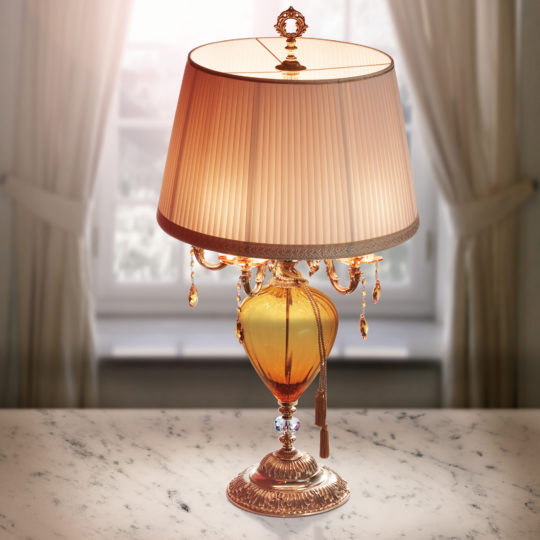 The Large Amber Glass Candelabra Style Table Lamp features a spacious amber-colored glass body with an elaborate gold base. Its cream-colored lampshade, embellished with a gold trim and hanging crystal accents, complements the design beautifully. Positioned on a marble surface, the lamp is framed by sheer curtains and a window in the background.