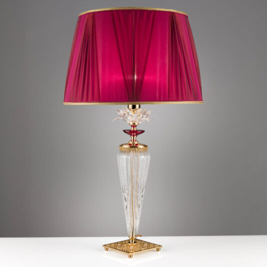The Luxury Table Lamp With Crystal Flower Detail boasts a tall, ornate glass base accented with gold and intricate designs. It features exquisite crystal flower details that add a touch of elegance. The vibrant magenta lampshade with a subtle sheen stands on a square base adorned with sophisticated patterns, all set against a plain gray background.