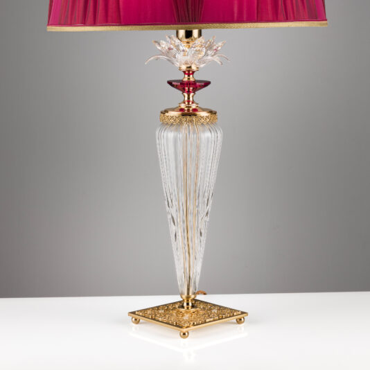 Luxury Table Lamp With Crystal Flower Detail The Luxury Table Lamp With Crystal Flower Detail features a gold base adorned with an intricate pattern and crystal flower accents. Its tall, tapered glass body showcases vertical grooves, complemented by a gold-colored accent near the top. The lamp is crowned with a red lampshade detailed with elegant pleating.
