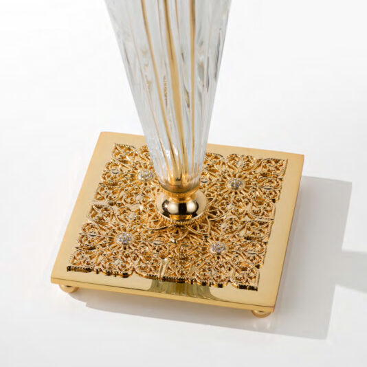 Luxury Table Lamp With Crystal Flower Detail A close-up of the Luxury Table Lamp With Crystal Flower Detail showcases its ornate base adorned with intricate gold filigree and floral patterns. The small embedded crystals add a touch of elegance to the square base, which supports a tall, tapered, and fluted transparent column.