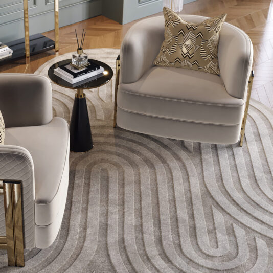 A stylish living room showcases two Modern Curved Armchairs in cream with gold accents and geometric patterned pillows. A small black round table between the chairs holds a reed diffuser. The floor is adorned with a gray and beige rug featuring an abstract wavy pattern.