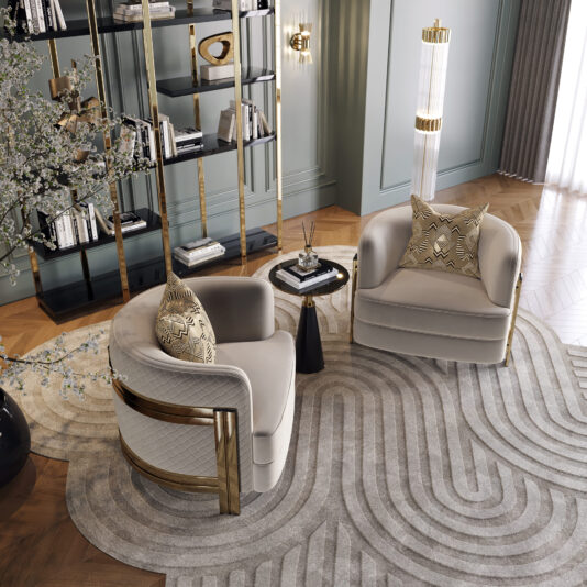 A stylish living room showcases two Modern Curved Armchairs adorned with patterned pillows, accompanied by a black and gold side table holding decorative items. The space is illuminated by a modern light fixture and features a bookshelf filled with books and décor pieces. The room is finished off with a beige patterned rug and wooden flooring.