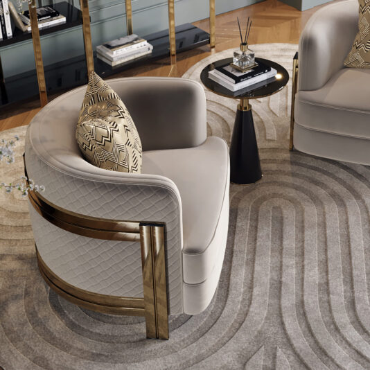 A luxurious seating area featuring the Modern Curved Armchair with plush, quilted upholstery and gold accents. Beside it, a stylish round side table holds books and a reed diffuser. The setup rests on an ornate, geometric-patterned rug, enhancing the contemporary decor.