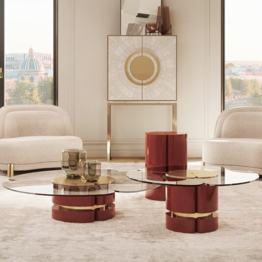 A contemporary living room showcasing a Modern Lacquered Coffee Table Set with red and gold bases, accompanied by beige upholstered chairs and a decorative console adorned with geometric artwork. Large windows allow natural light to flood the space while offering a stunning view of the cityscape in the background.