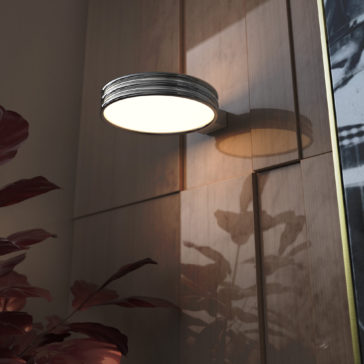 Modern Spot Style Wall Light A Modern Spot Style Wall Light with a round, illuminated shade is mounted on a wooden paneled wall. Red-leaved plants in the foreground cast shadows, enhancing the room's warm and artistic ambiance.