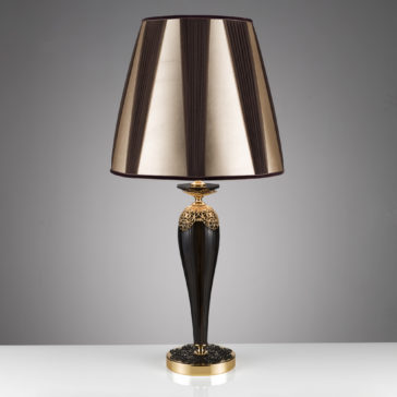 Ornate Black And Gold Glass Table Lamp The Ornate Black And Gold Glass Table Lamp features a sleek, modern design with a tall, black, tapered base adorned with intricate gold accents. It is topped with a large, cone-shaped lampshade that boasts gold and black vertical stripes, set against a plain gray background.
