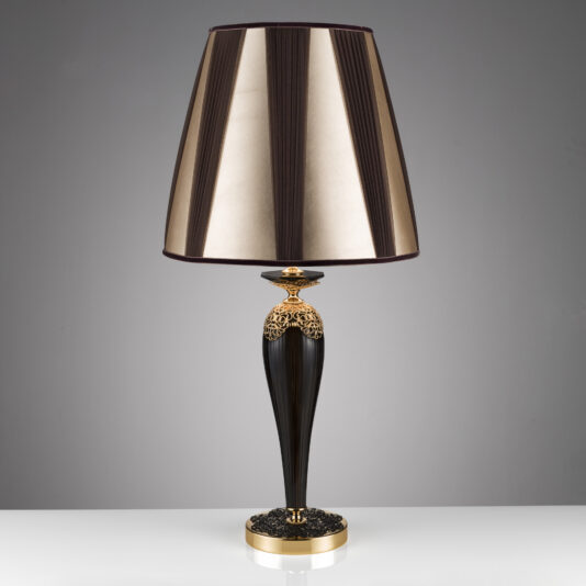 The Ornate Black And Gold Glass Table Lamp features a sleek, modern design with a tall, black, tapered base adorned with intricate gold accents. It is topped with a large, cone-shaped lampshade that boasts gold and black vertical stripes, set against a plain gray background.