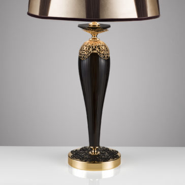Ornate Black And Gold Glass Table Lamp The Ornate Black And Gold Glass Table Lamp exudes sophistication with its elegant black and gold design. It features a tall, tapered base adorned with intricate filigree details near the top and bottom, complemented by a wide, gold drum-shaped lampshade. This luxurious lamp is beautifully displayed on a white surface against a neutral background.