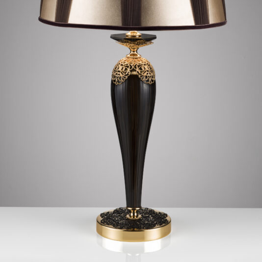 The Ornate Black And Gold Glass Table Lamp exudes sophistication with its elegant black and gold design. It features a tall, tapered base adorned with intricate filigree details near the top and bottom, complemented by a wide, gold drum-shaped lampshade. This luxurious lamp is beautifully displayed on a white surface against a neutral background.