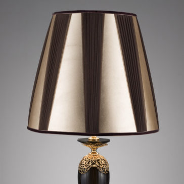 Ornate Black And Gold Glass Table Lamp A table lamp with a sophisticated, gold-toned base and an elegant black body. The lampshade features vertical stripes in alternating shades of brown and gold, adding to the stylish look of this Ornate Black And Gold Glass Table Lamp. The background is plain and gray.