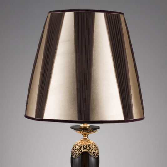 A table lamp with a sophisticated, gold-toned base and an elegant black body. The lampshade features vertical stripes in alternating shades of brown and gold, adding to the stylish look of this Ornate Black And Gold Glass Table Lamp. The background is plain and gray.