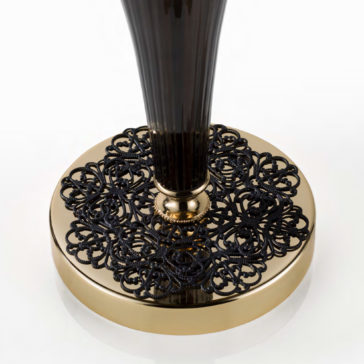 Ornate Black And Gold Glass Table Lamp The Ornate Black And Gold Glass Table Lamp features a tall, slender black body set atop an opulent gold base intricately adorned with black lace-like patterns. This luxurious and sophisticated design is enhanced by the white background, which highlights the lamp's exquisite details and craftsmanship.