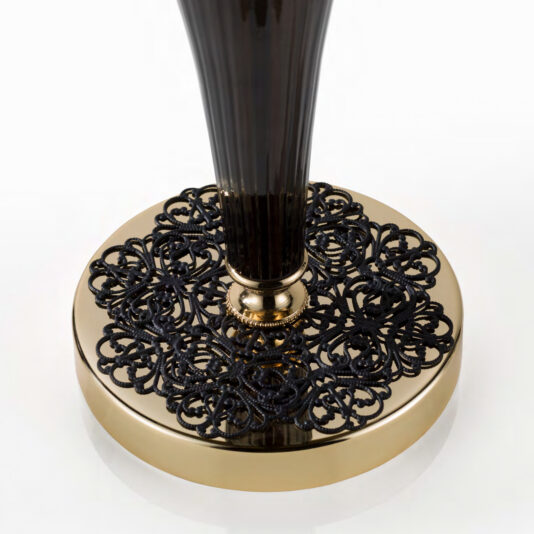The Ornate Black And Gold Glass Table Lamp features a tall, slender black body set atop an opulent gold base intricately adorned with black lace-like patterns. This luxurious and sophisticated design is enhanced by the white background, which highlights the lamp's exquisite details and craftsmanship.