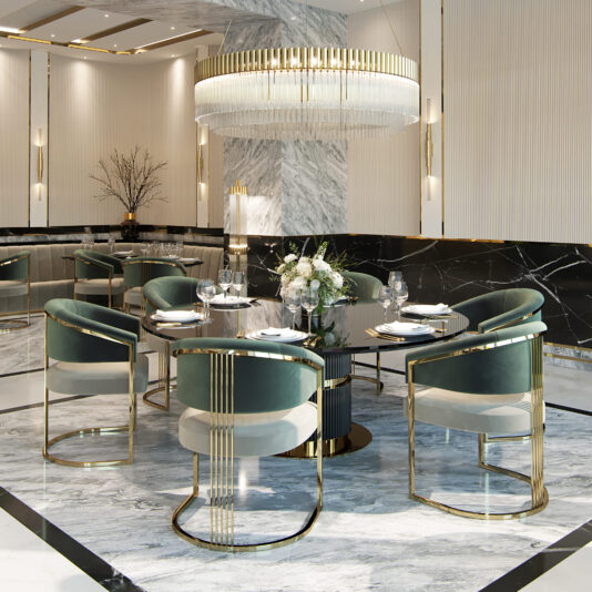 Two-Tone Art Deco Style Curved Dining Chair A luxurious dining room with a round black table set for six, surrounded by Two-Tone Art Deco Style Curved Dining Chairs in green and gold. A large, elegant chandelier with crystal accents hangs above the table. The room features marble flooring, modern art decor, and a vase with white flowers as a centerpiece.