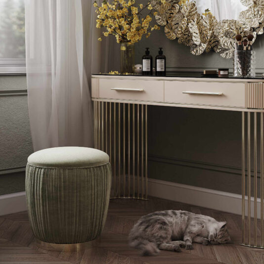 A chic vanity setup features a sleek desk with gold accents, a Velvet Pouffe With Gold Plated Base, elegant toiletries, a flower vase, and a leaf-shaped wall mirror. A gray cat lies comfortably on the wooden floor next to the pouffe as sheer curtains filter natural light.