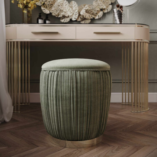 A Velvet Pouffe With Gold Plated Base sits elegantly in front of a modern vanity table adorned with gold accents. The table features two drawers and is complemented by a decorative mirror above it. Wooden herringbone flooring, along with a floral arrangement, adds warmth to the stylish room.