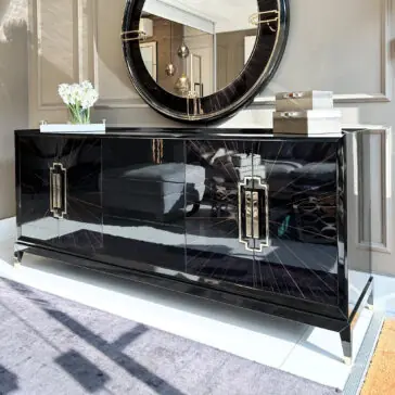 High End Veneer Art Deco Style Sideboard A High End Veneer Art Deco Style Sideboard in glossy black enhances the room with its modern design. Positioned above it is a round mirror, while decorative items and a vase of white flowers adorn its top. The neutral walls and gray rug contribute to the room's sophisticated ambiance.