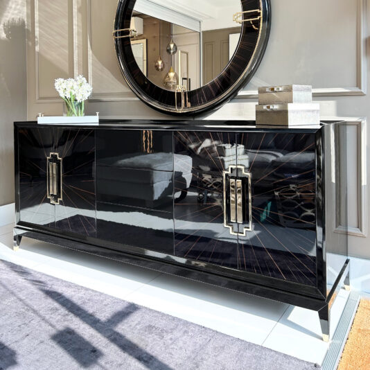 A High End Veneer Art Deco Style Sideboard in glossy black enhances the room with its modern design. Positioned above it is a round mirror, while decorative items and a vase of white flowers adorn its top. The neutral walls and gray rug contribute to the room's sophisticated ambiance.