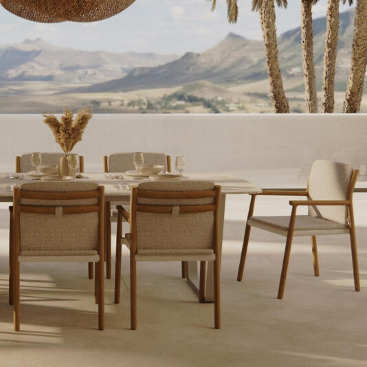 A Contemporary Outdoor Dining Set With Teak Chairs featuring a wooden table is beautifully arranged on a terrace. The set includes six cushioned seats, complemented by plates, glasses, and a vase of dried flowers. The scene is enhanced by tall palm trees and distant mountains serving as an idyllic backdrop to this modern setup.