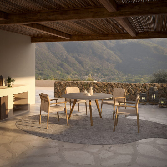 A cozy outdoor patio features a wooden dining table paired with four Contemporary Outdoor Teak Dining Chairs, all set on a stone surface. Overhead, a wooden pergola provides soothing shade. A stone wall and scenic mountain view enhance the tranquil ambiance, while decorative items are arranged on a built-in counter to the left.