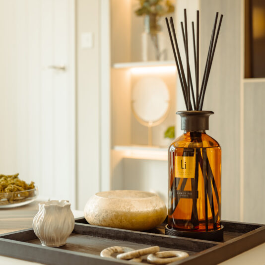In a well-lit room, an Extra Large Luxury Amber Fie Room Diffuser is elegantly displayed on a black tray alongside a round beige bowl, a white textured vase, and intertwined stone rings. The background features a light-colored shelf accented with soft lighting.