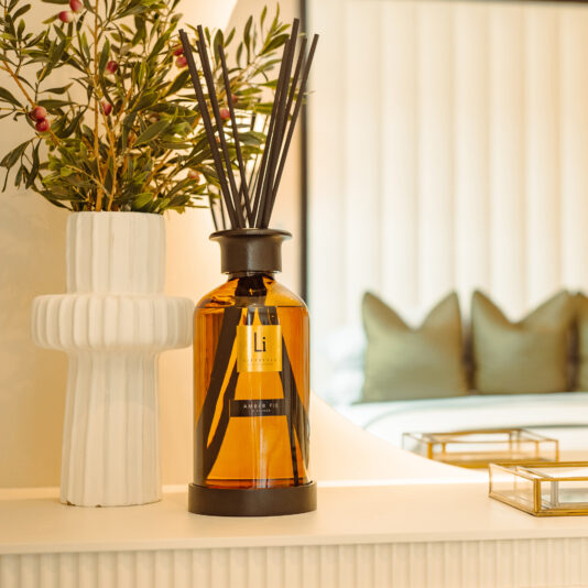 An Extra Large Luxury Amber Fie Room Diffuser with aromatic fragrance sticks sits on a white countertop, accompanied by a ceramic vase containing green and red plant branches. In the background, a bed featuring a white padded headboard and green pillows can be partially seen.