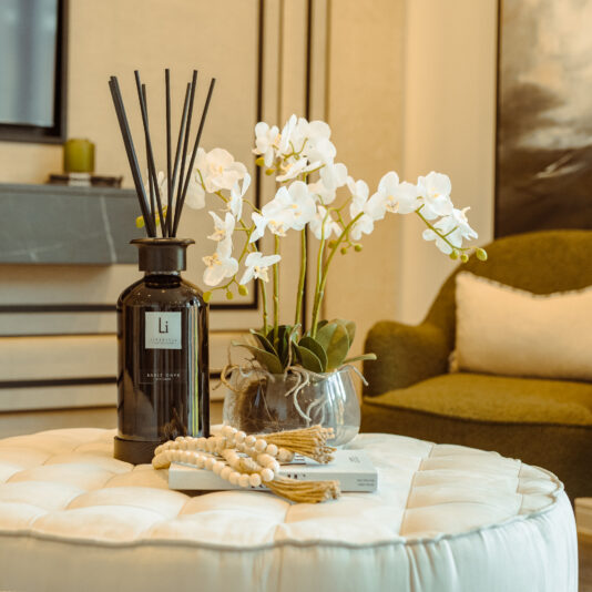 A cozy living room scene with a round, tufted ottoman holding an Extra Large Luxury Sable Onyx Room Diffuser, a small vase of white orchids, and a decorative bead string. In the background, there is a green armchair with a white pillow and framed artwork on the wall.